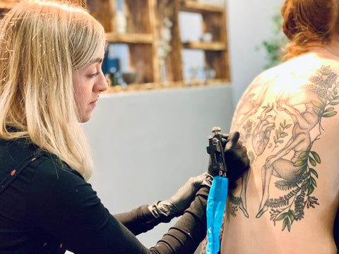 Frances is sitting down and tattooing a large design on a client's back.