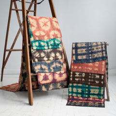 Ta'na'na silk raffia table runners handmade in Madagascar displayed on a tall ladder and a step ladder. All colors are layered: indigo, brown, charcoal, burgundy, terra cotta, and turquoise. Two different patterns are shown: turtle pattern and cocoon and moth pattern.