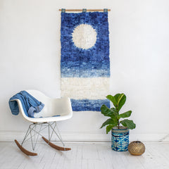 image showing the full moon reflecting on water wall hanging, hung on a plane white wall. in front of the artwork is a white rocking chair with a blue denim jacket draped over the back of it. beside the chair, also in front of the wall hanging, is a ta'na'na silk sapphire blue wild silk basket with a houseplant in it. The artwork itself is in a bold shade of blue with a gradient, surrounding a round white section representing the moon. It is handmade in Madagascar. there is a white stripe towards the bottom representing the moon's reflection in the water.
