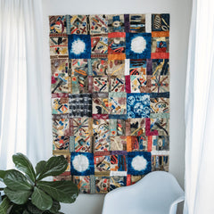 Madagascar Dreaming wall hanging, a large, delightful collage filled with more and more detail the closer you look, hangs on a white wall between two windows covered in white curtains. In from of the wall hanging is a white chair and a houseplant.