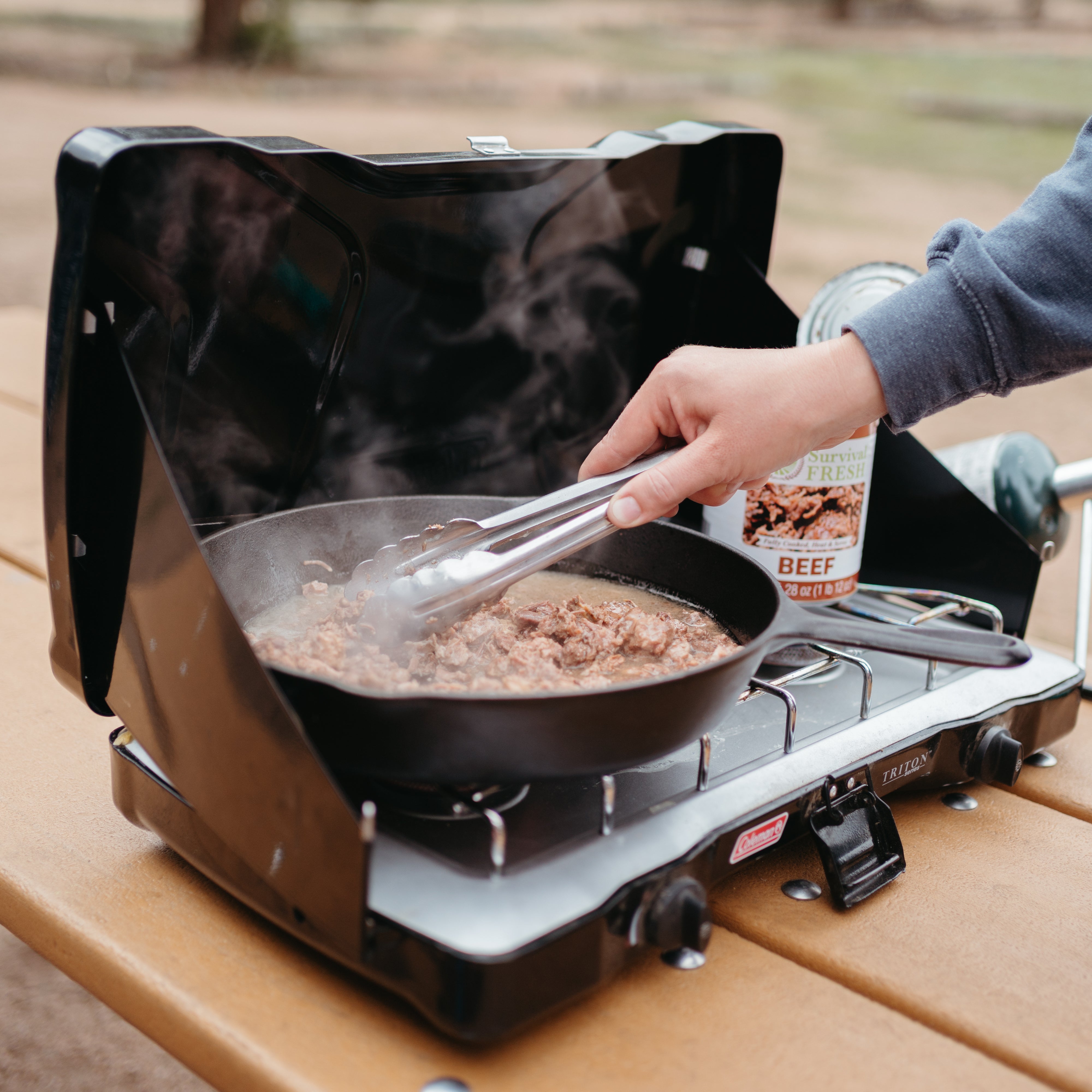 Person cooking beef on a portable gas stove outdoors.