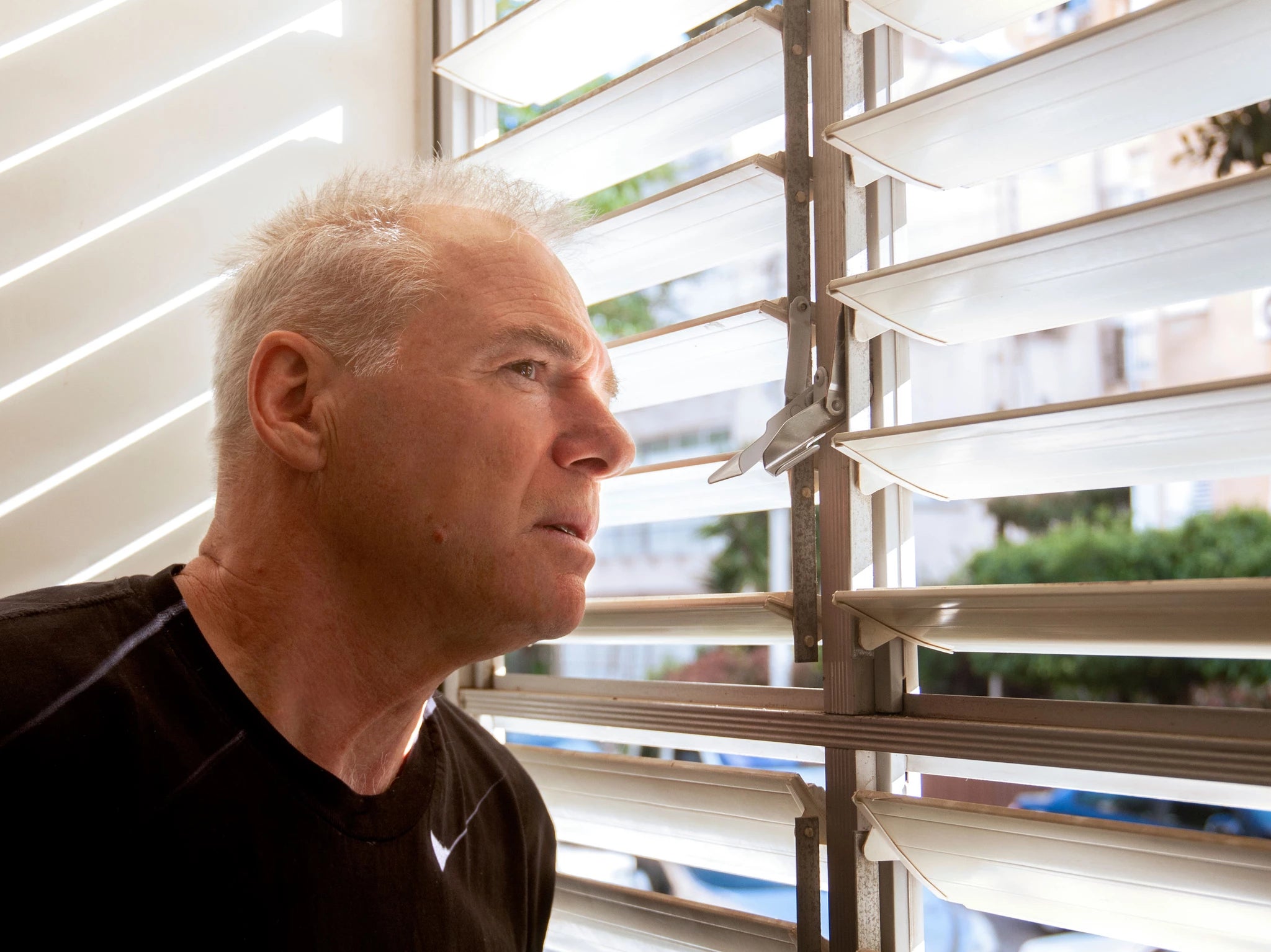 Man looking outside through venetian blinds with a contemplative expression.