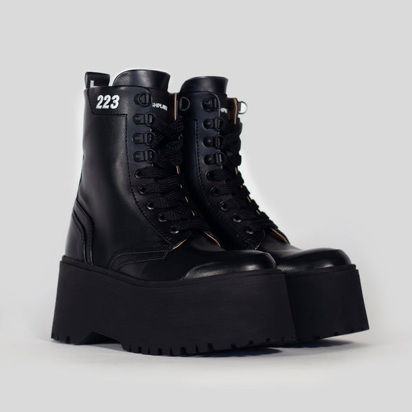 The chunky platform sole RIOT! boot which is 100% vegan, and made from recycled and upcycled food waste - it features a black apple leather upper and corn leather lining. The boots sit on an angle against a light grey background and feature black laces, rubber label, and 223 embroidery in white.