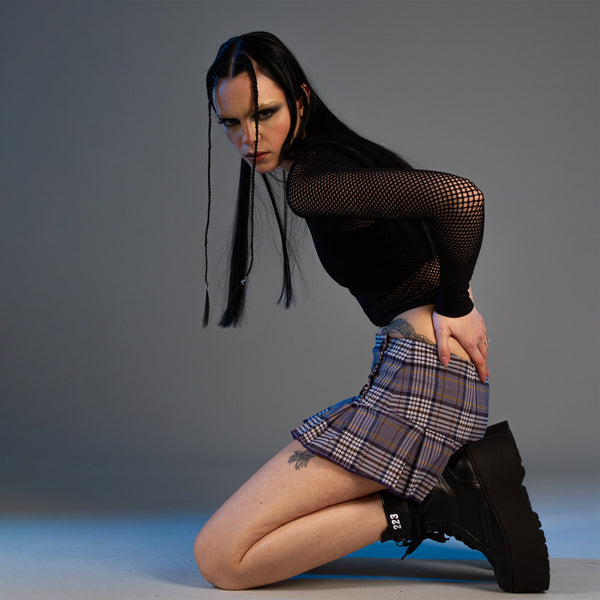 A girl wearing a netted mesh top in black, and a pleated mini tartan kilt skirt, wears chunky black huge platform goth style vegan boots made from food waste like apple leather and corn leather sits on her knees leaning forward with her hands on her back directly looking into the camera.