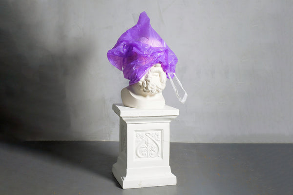 A white sculputre of a man's face on a pedestal is covered by a violet plastic bag