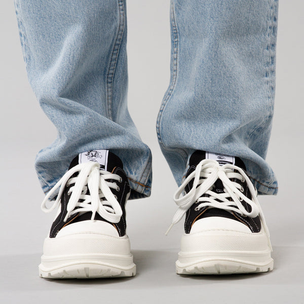 The front view of Trash Planet's CARTER 223 sneaker, which has a 90's punk aesthetic, and a chunky sole with rubber toe cap. The mesh is black, and made from 100% recycled ocean plastic. The model is wearing blue jeans which sit over the top of the sneakers.