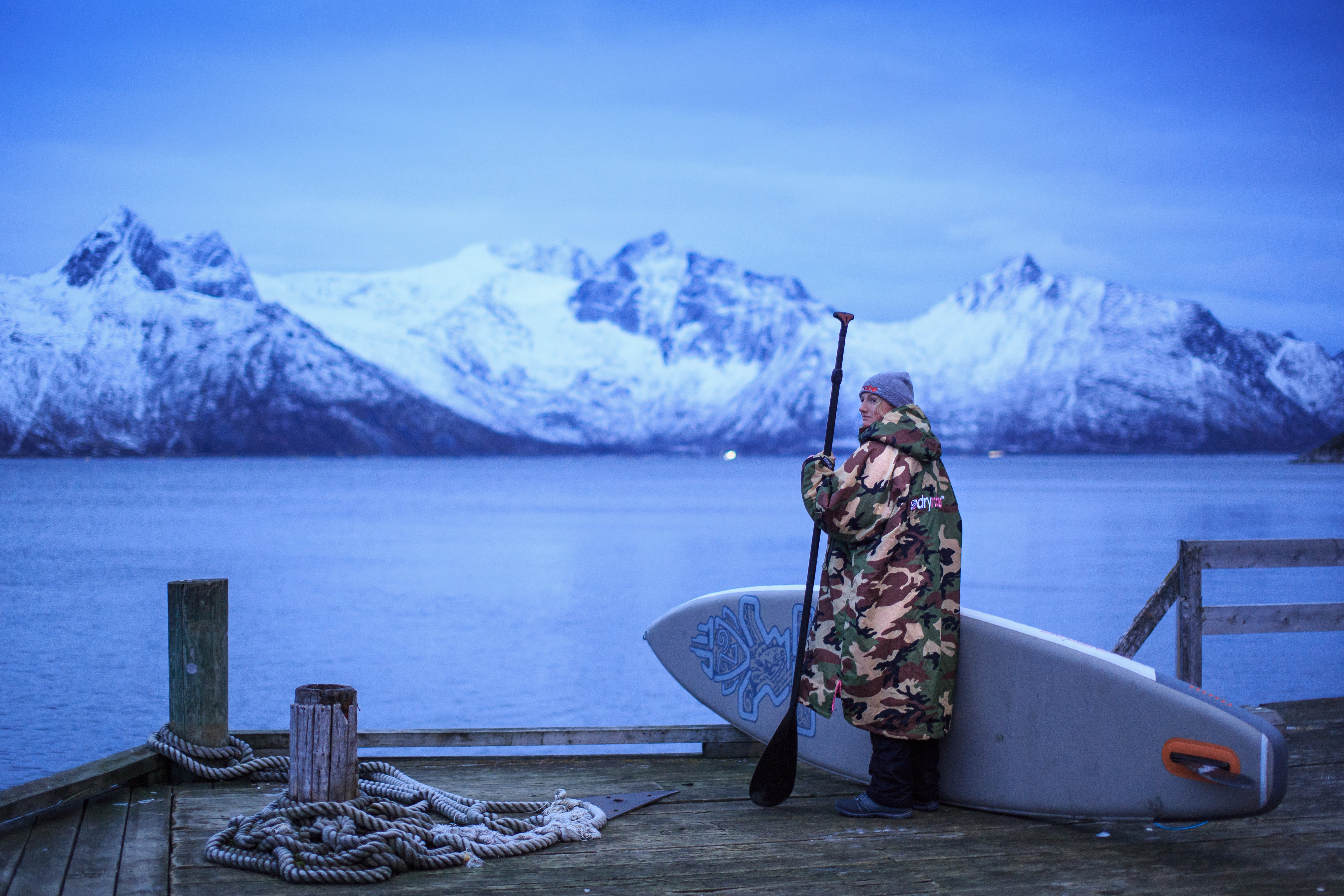 Cal Major standing by a Fjord in Norway in a dryrobe, holding a stand up paddleboard