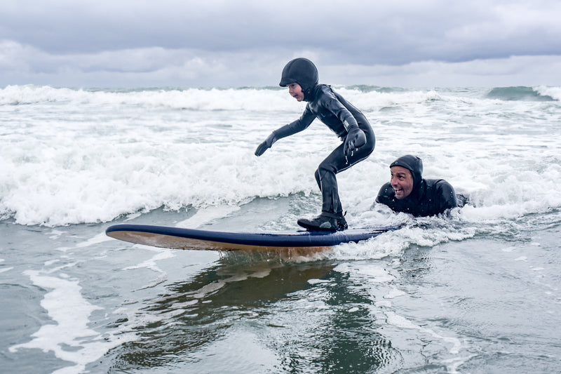 A kid in a hooded wetsuit catching a wave with a surf instructor behind him 