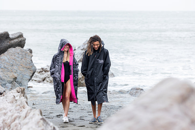 A woman and man in swim gear and dryrobe walking on the beach smiling 