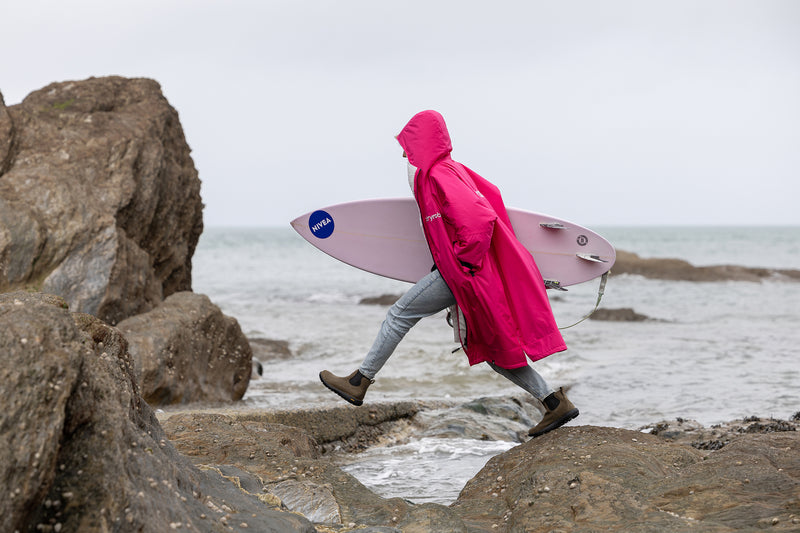 A woman wearing a bright pink dryrobe holding a surfboard and walking over rocks by the beach