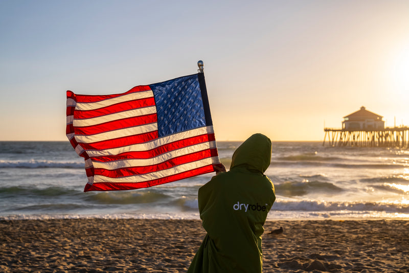 A person stood in a forest green dryrobe holding an American flag on the beach by the sea