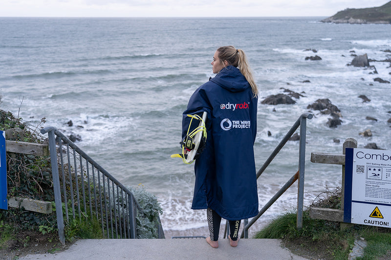 Female surfer holding a surfboard and wearing a Wave Project dryrobe Advance by the sea