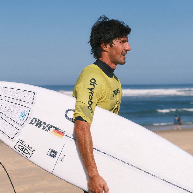 A male surfer wearing a yellow competition rash vest walking away from the sea holding his surfboard
