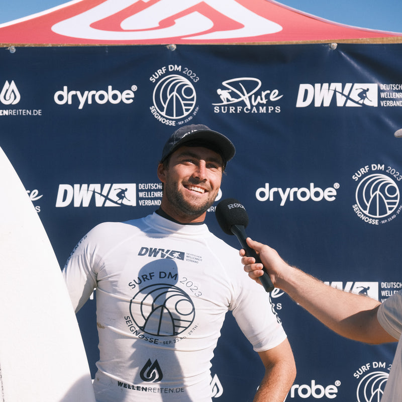 A male surfer being interview wearing a rash vest and holding a surfboard