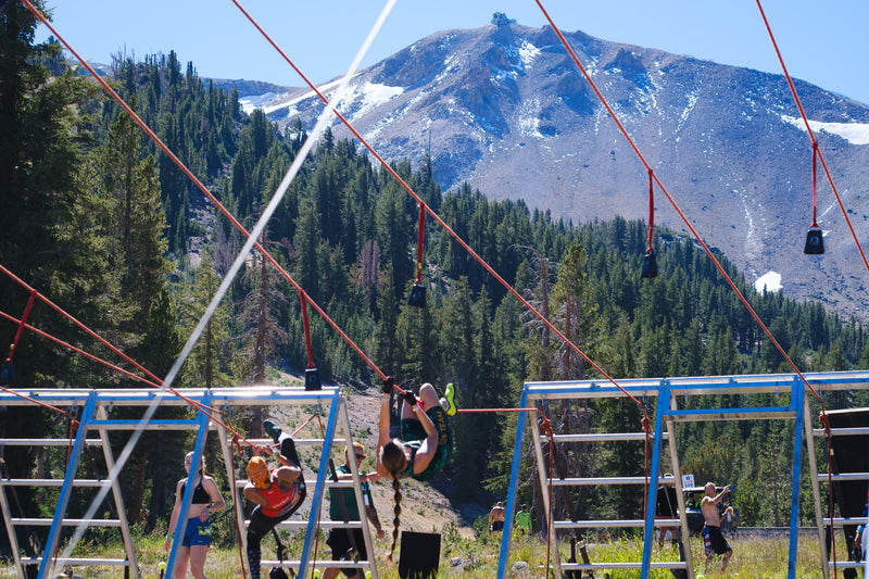 Two women competing on a rope obstacle with trees and a mountain in the background