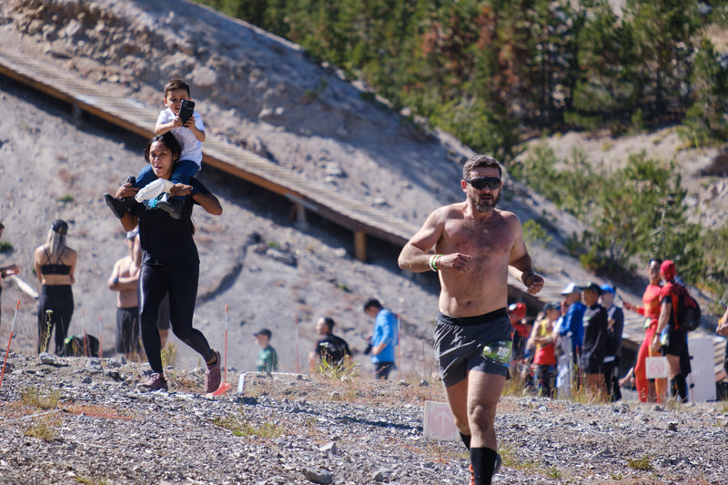Man running with his shirt off followed by woman with a child on her shoulders