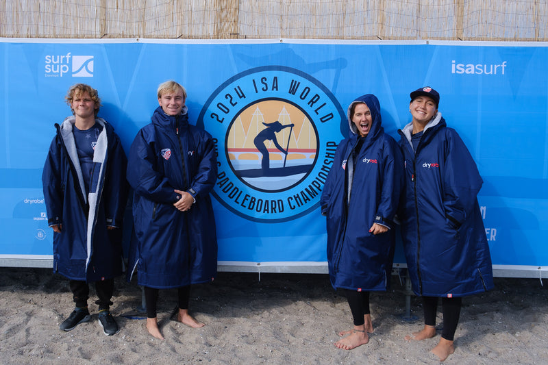 Team USA in branded dryrobes smiling next to the 2024 ISA World SUP and Paddleboard Championship sign