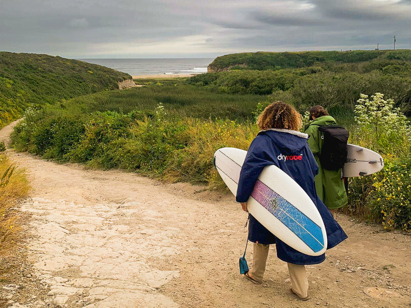 Two surfers walking to the beach carrying board and wearing dryrobes