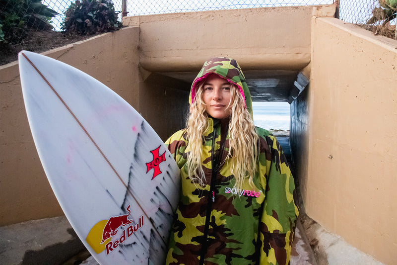 Izzi Gomez holding her surfboard in front of a tunnel leading to the beach, whilst wearing a camo dryrobe Advance