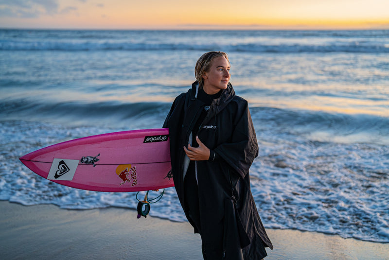 Izzi Gomez holding a surfboard wearing a wetsuit and dryrobe Lite on a beach at sunset