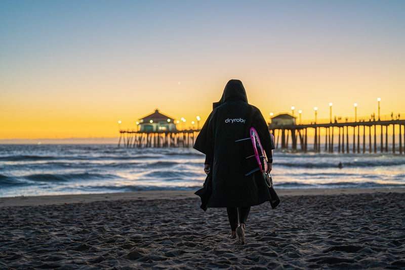 A person wearing a dryrobe Lite and carrying a pink surfboard to the seashore