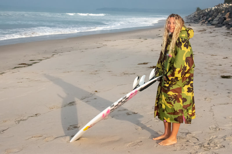 Izzi Gomez on the beach holding her surfboard wearing a camo dryrobe Advance