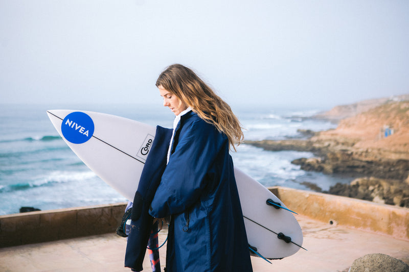 Lucy Campbell holding surfboard by the shore in a dryrobe 