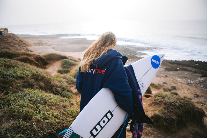 Lucy Campbell wearing a dryrobe and holding a surfboard walking over the sand dunes to the sea