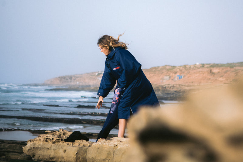 Female surfer Lucy Campbell on the beach putting her wetsuit on while wearing a navy dryrobe
