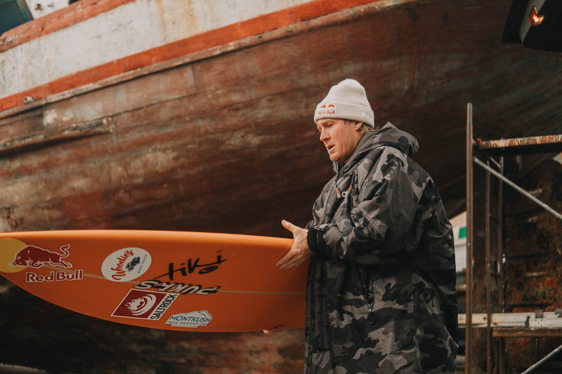 Andrew Cotton holding a surfboard and wearing a dryrobe