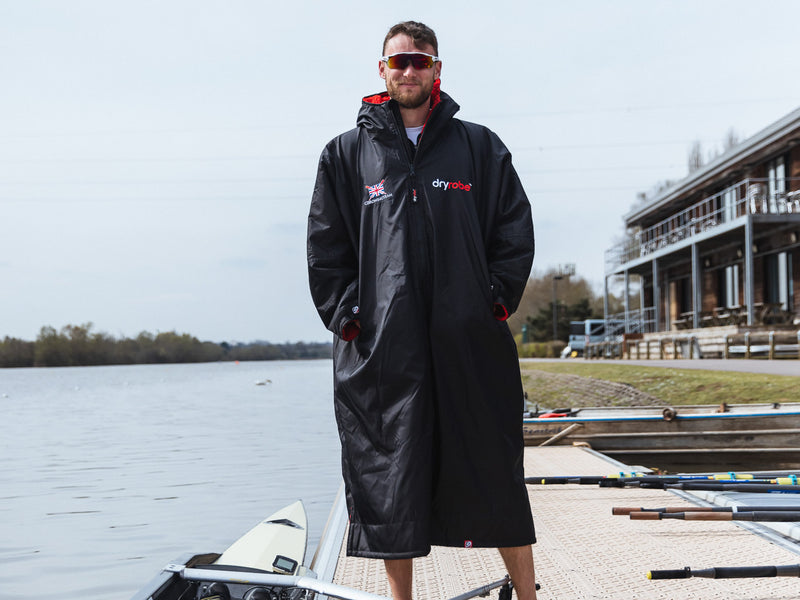 A British rower stood by water wearing a dryrobe 