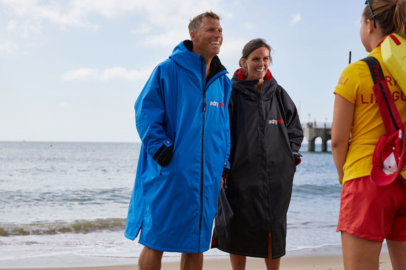 Two people stood in dryrobes talking to a lifegaurd on the beach and smiling 