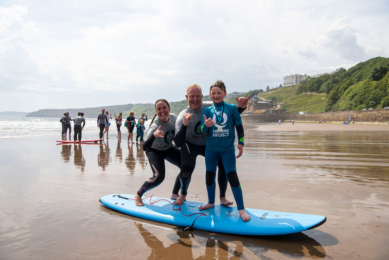 Kids and volunteers stood on a foam surfboard on the beach at a Wave Project session