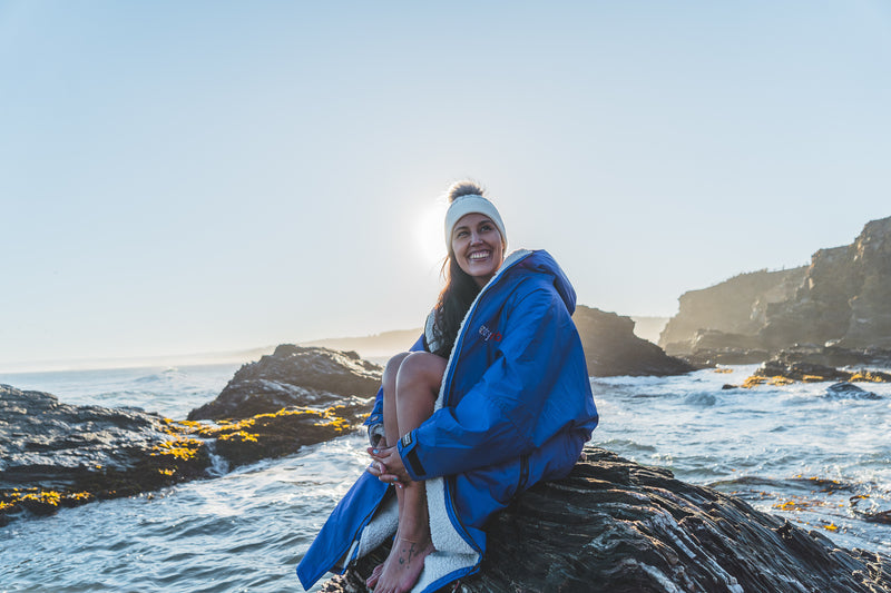 Bárbara Hernández Huerta sitting on rocks by the sea wearing a dryrobe changing robe and a bobble hat