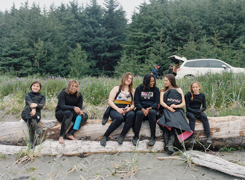 A group of kids sat on a log outside near a forest wearing wetsuits