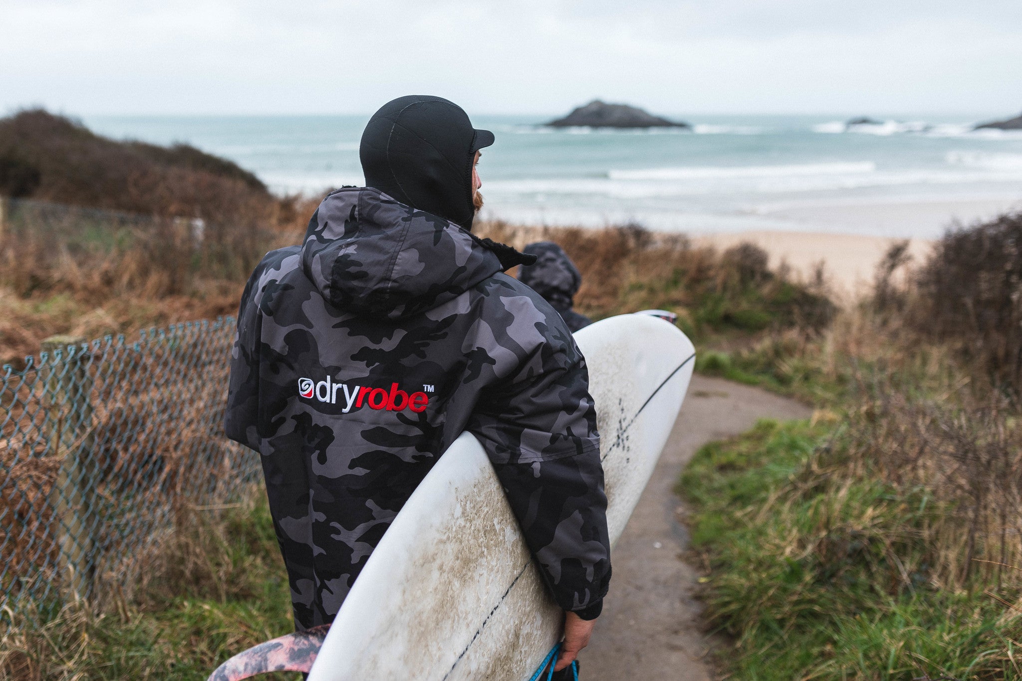 Ben skinner carry his surfboard to the beach whilst wear a Camo Black dryrobe Advance