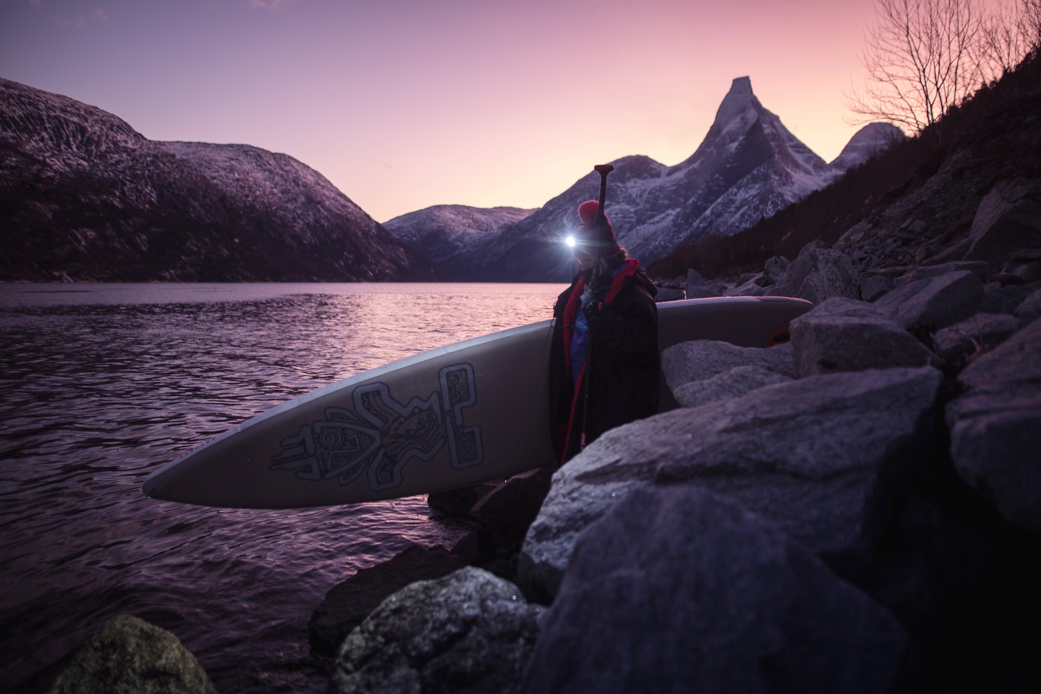 Cal Major carrying her SUP to the edge of a fjord in the dusk