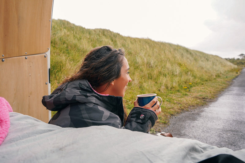 Woman sat in the back of a camper van drinking a coffee and looking out onto the road