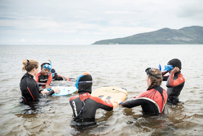 Cal Major knelt in the sea with a group children wearing wetsuits and snorkels looking at marine life 