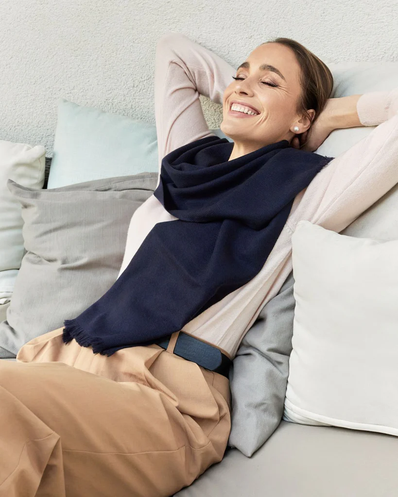 Smiling woman reclines on a sofa with her arms behind her head, wearing a cashmere scarf and a cashmere jumper.