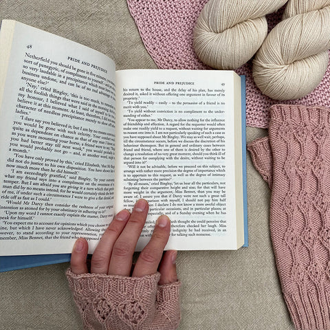 Pride and prejudice book open held by left hand wearing knitted fingerless mitts. Flat lay with other mitt to the right and two skeins of yarn above. 