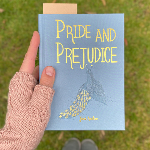 Left hand wearing a pink knitted fingerless mitt, holding a blue Pride and Prejudice book