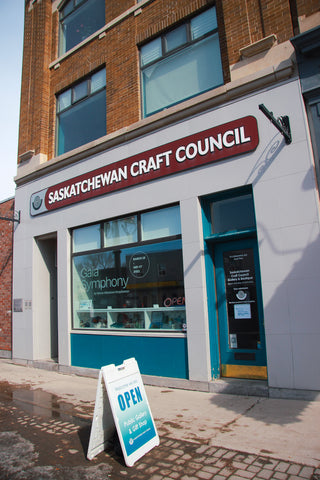 A photo of the Saskatchewan Craft Council building storefront. There is a large window that displays a vinyl wordmark reading "Gaia Symphony", and a display of craft in the bottom portion of the windowbox. 