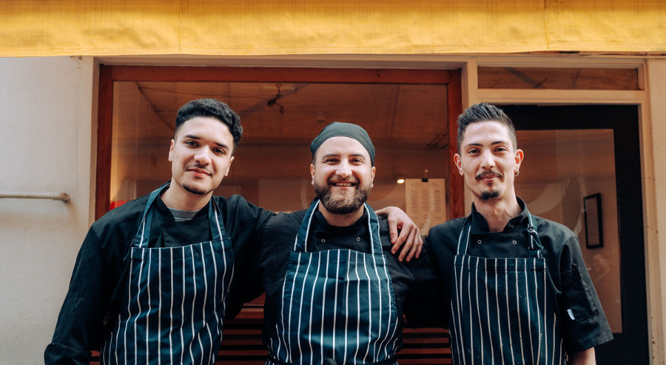 Three men in black chef's hats, aprons, and black polo shirts stand smiling with their arms around each other in front of a building with yellow awning.