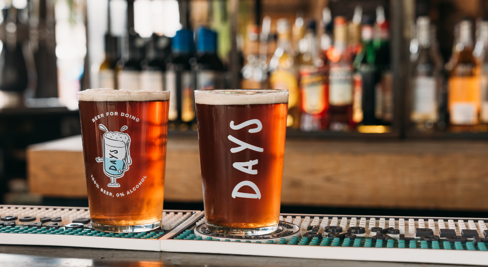 Two pint glasses full of Days beer and branded with the white Days logo are placed on a bar counter. Wine bottles and bar equipment are out of focus in the background.