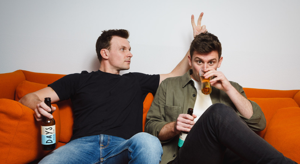 Two white men with brown hair sit on an orange sofa. One wearing a black t-shirt holds a peace sign behind the others head, as the other wearing a white t-shirt and green shirt sips beer from a glass.