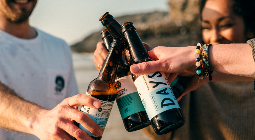 Three people clink their Days beer bottles together as they stand on a beach.