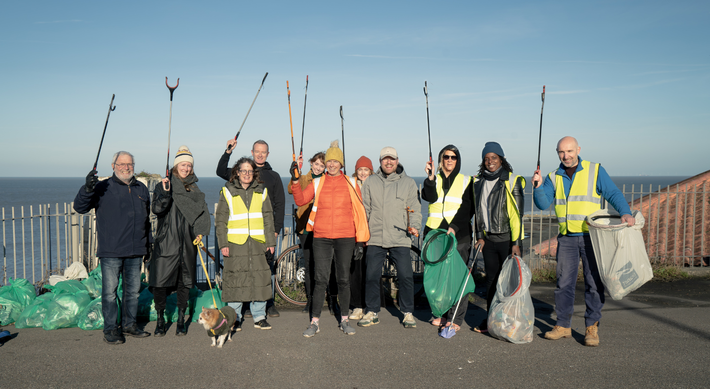 A group of people holding litter pickers and plastic rubbish bags, smile at the camera. To the far left, there are full green rubbish bags and behind them is the sea and a blue sky.