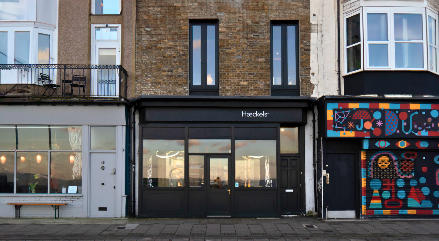 The storefront of Haeckels in Margate, which is painted in all black, sandwiched between other shops on the street. The sunset sky is reflected on the windows.