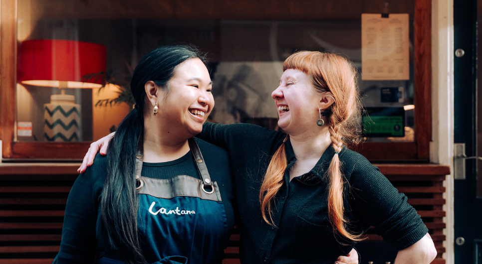 Two smiling women, one with black hair wearing an apron with the Lantana logo and the other with ginger hair wearing a black polo shirt and orange bottoms, stand side by side with their arms around each other in front of a shop front.